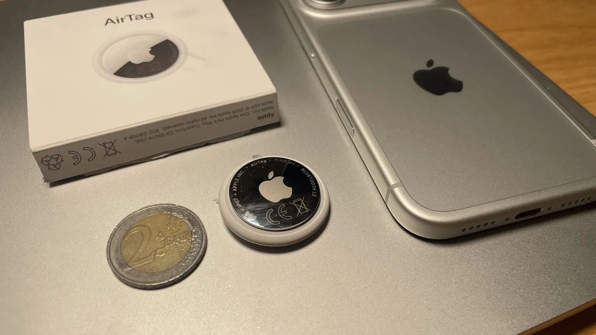 Apple AirTag photographed beside a two-euro coin, an iPhone, and its retail box to show its compact size